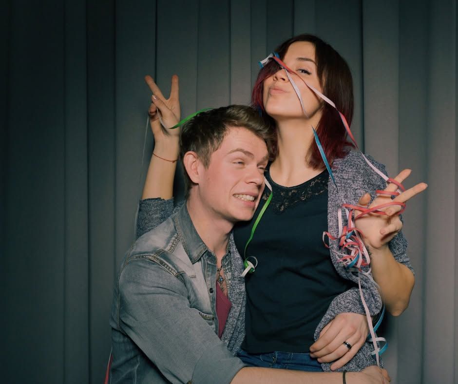 Two people posing together indoors, smiling and making playful hand gestures.