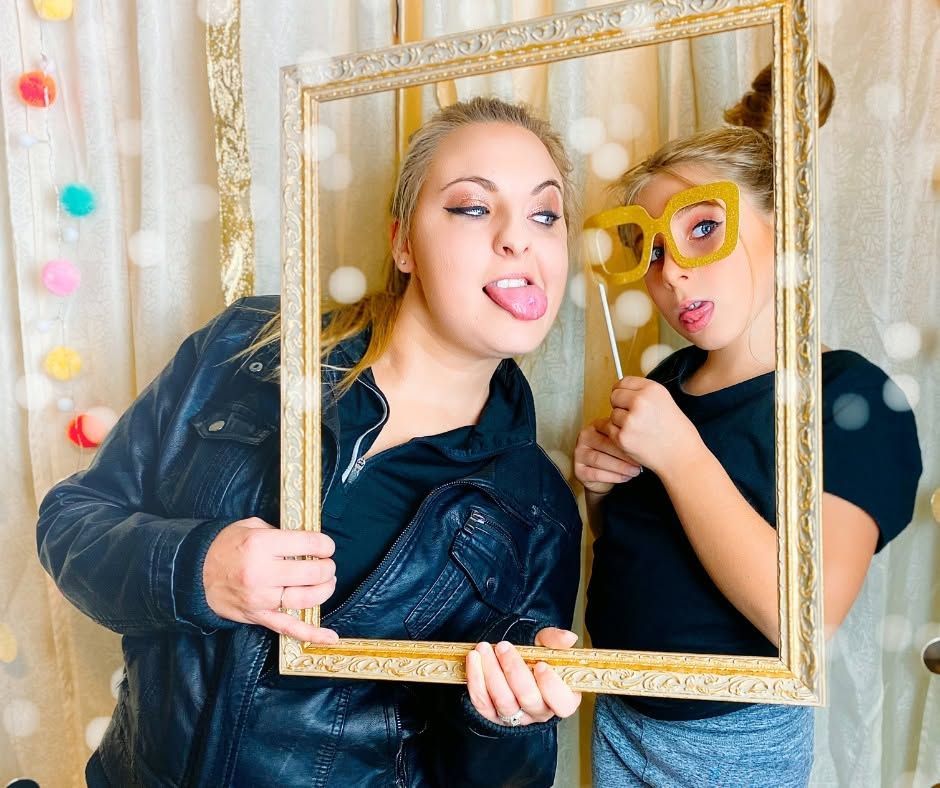 Two people posing with a gold picture frame, one wearing novelty glasses, against a white curtain backdrop