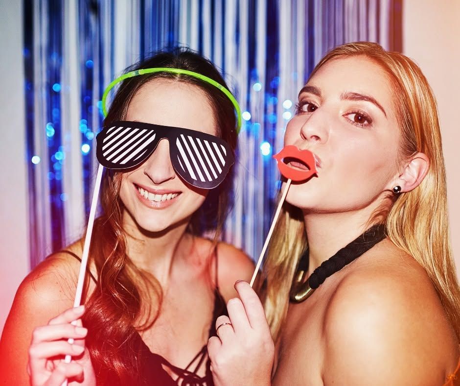 Two smiling partygoers posing with props, one in striped sunglasses and one wearing red lips, against a shiny backdrop