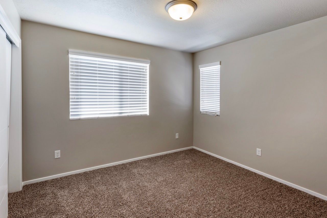 Empty beige bedroom with two windows, white blinds, and brown carpet.