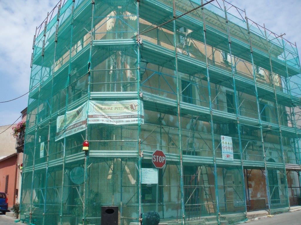 Edificio in fase di ristrutturazione, coperto da impalcature verdi e reti di sicurezza. Cartello stradale visibile.