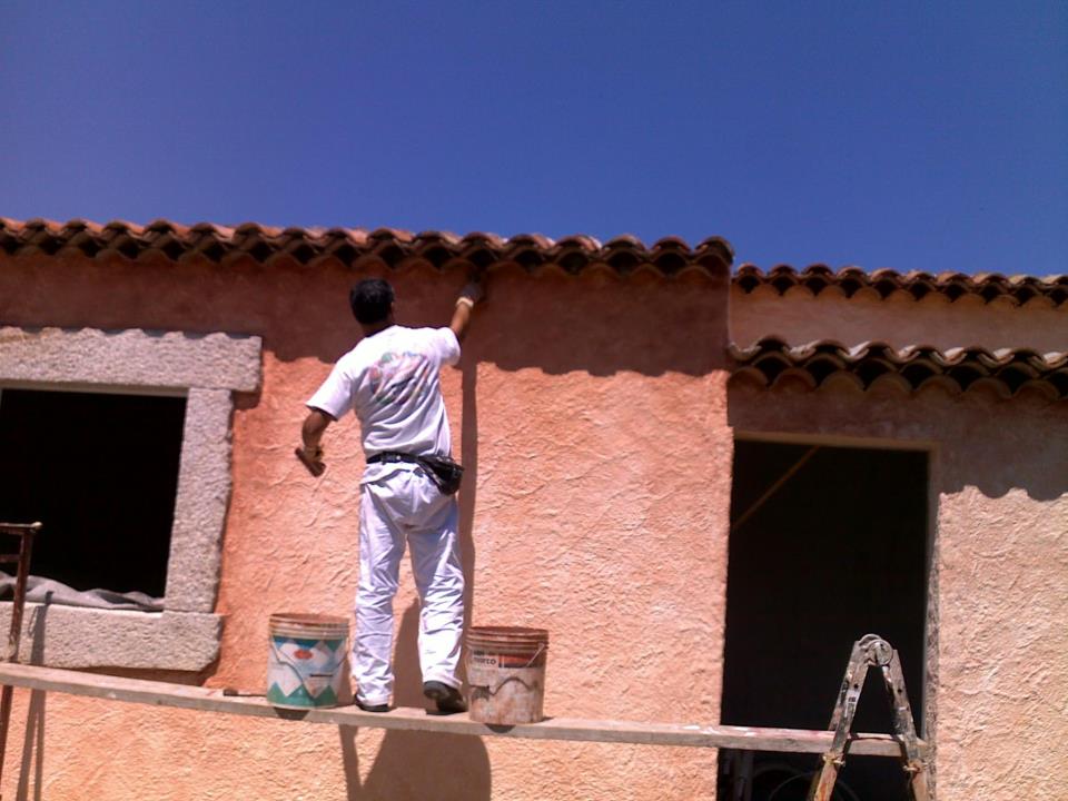 Persona che dipinge un edificio in stucco con pareti rosa sotto un cielo azzurro, utilizzando una scala e assi di legno.