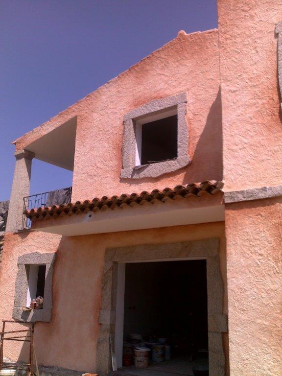 Edificio con stucco color salmone, infissi in pietra e un balcone sotto un cielo azzurro.