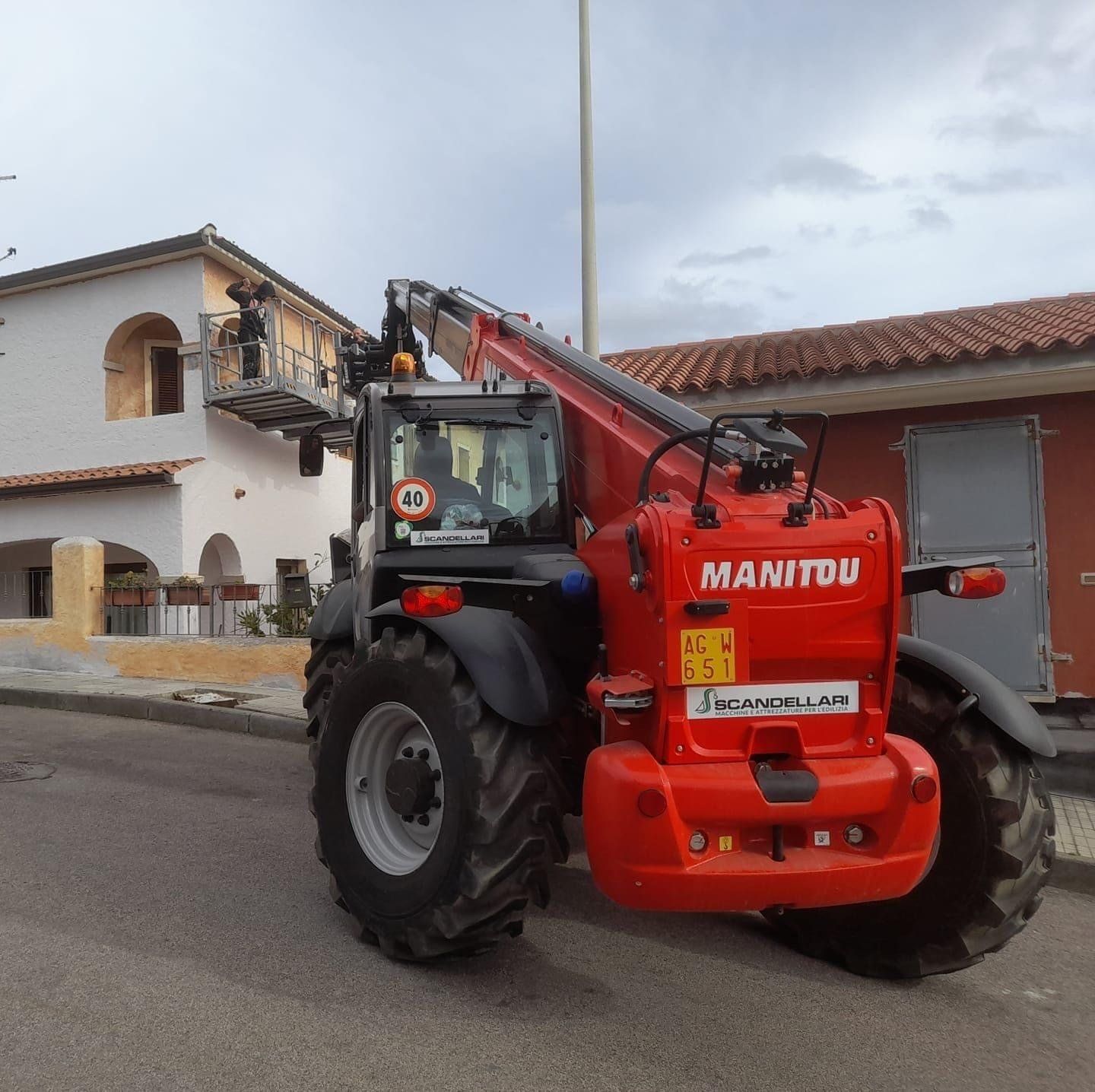 Sollevatore telescopico Manitou rosso con piattaforma di lavoro vicino a un edificio.