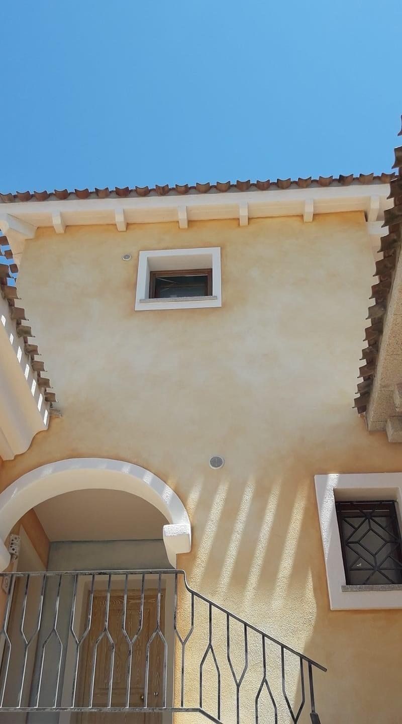 Esterno di un edificio con pareti in stucco giallo chiaro, piccole finestre e un tetto in terracotta contro un cielo azzurro.