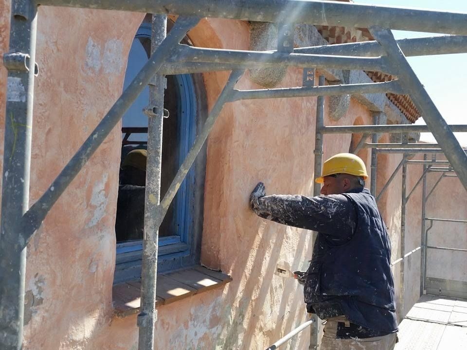 Un uomo con il casco applica lo stucco al muro di un edificio con una finestra blu. È visibile un'impalcatura.