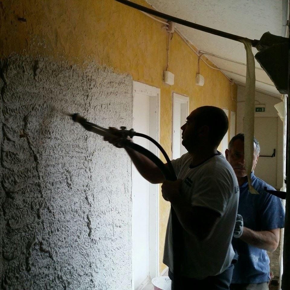 Uomo che spruzza stucco testurizzato sulla facciata di un edificio. Una seconda persona lo aiuta. Sfondo giallo.
