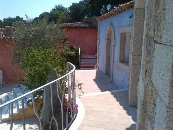 Percorso tra gli edifici, uno blu e uno rosso, con porta ad arco, muro in pietra e ringhiera in metallo.