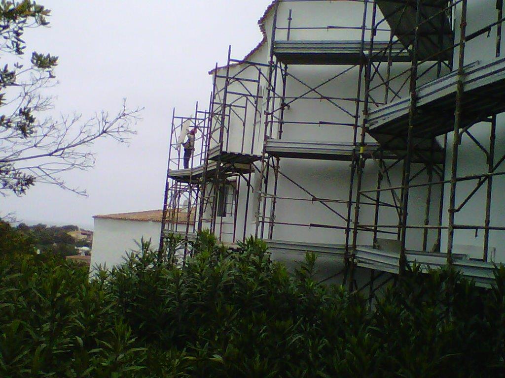 Esterno di un edificio con impalcature; operaio sulla piattaforma, cielo nuvoloso, cespugli verdi in primo piano.