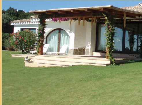 Un edificio in stucco bianco con porte ad arco e una veranda in legno, circondato da erba verde e piante.