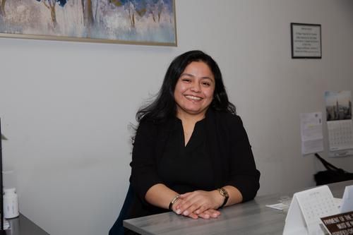 Smiling woman seated at an office desk with a laptop and paperwork, wearing a black blouse.