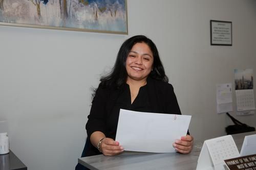 Smiling woman in a black blazer seated at a desk, holding a document in an office.