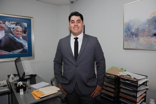 Man in a gray suit standing in an office beside a desk, framed photos, and stacked files
