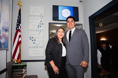 Two people pose indoors beside an American flag and wall sign, with a TV screen in the background.