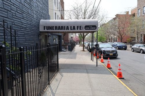 Sidewalk outside a funeral home with a black awning, parked cars, and orange traffic cones on the street.