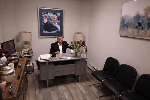 Person seated behind a desk in a small office, with black chairs, framed art, and a lamp.
