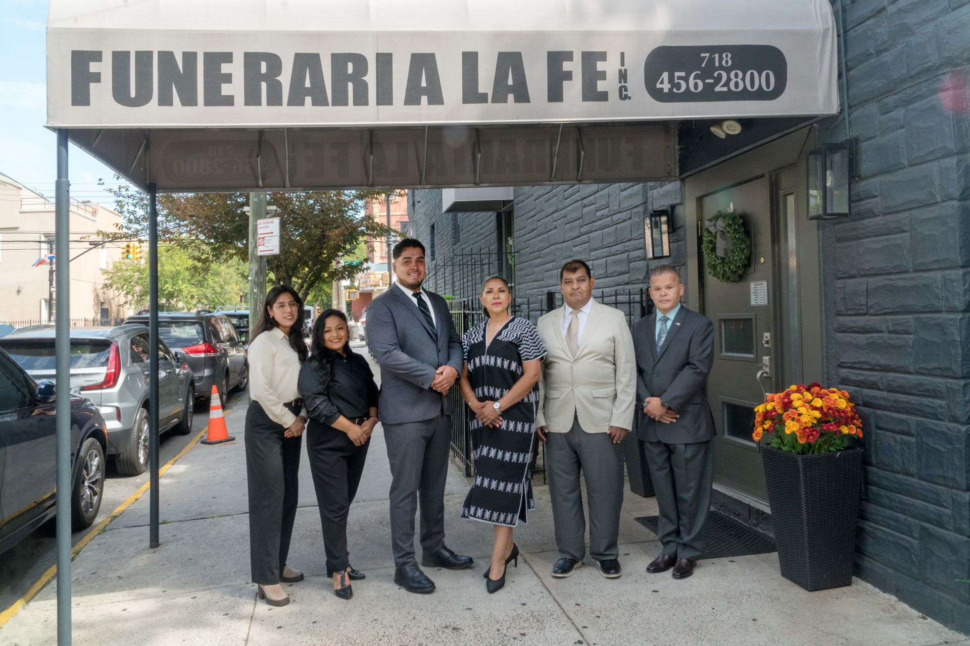 Six people standing outside a funeral home entrance with a sign and flower arrangement.