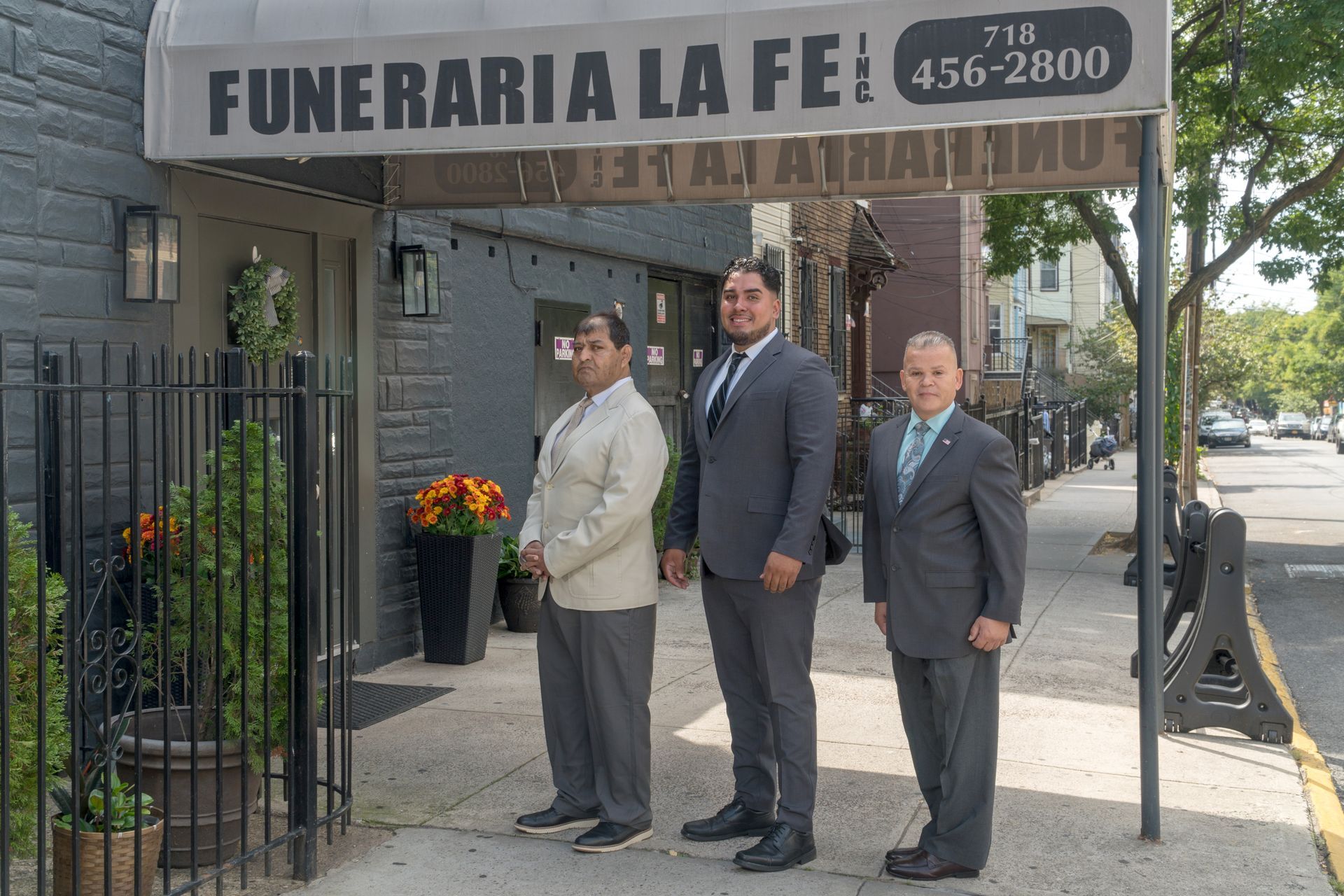 Three men standing outside Funeraria La Fe under a black awning, near the entrance and flower arrangements.