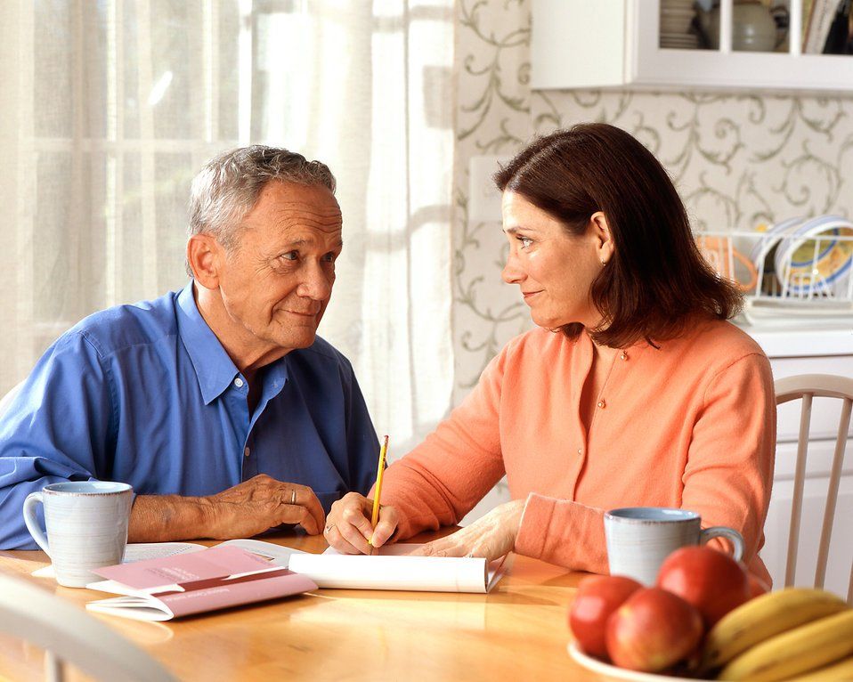Two people talking at a kitchen table with notebooks, mugs, and fruit in a bright home kitchen