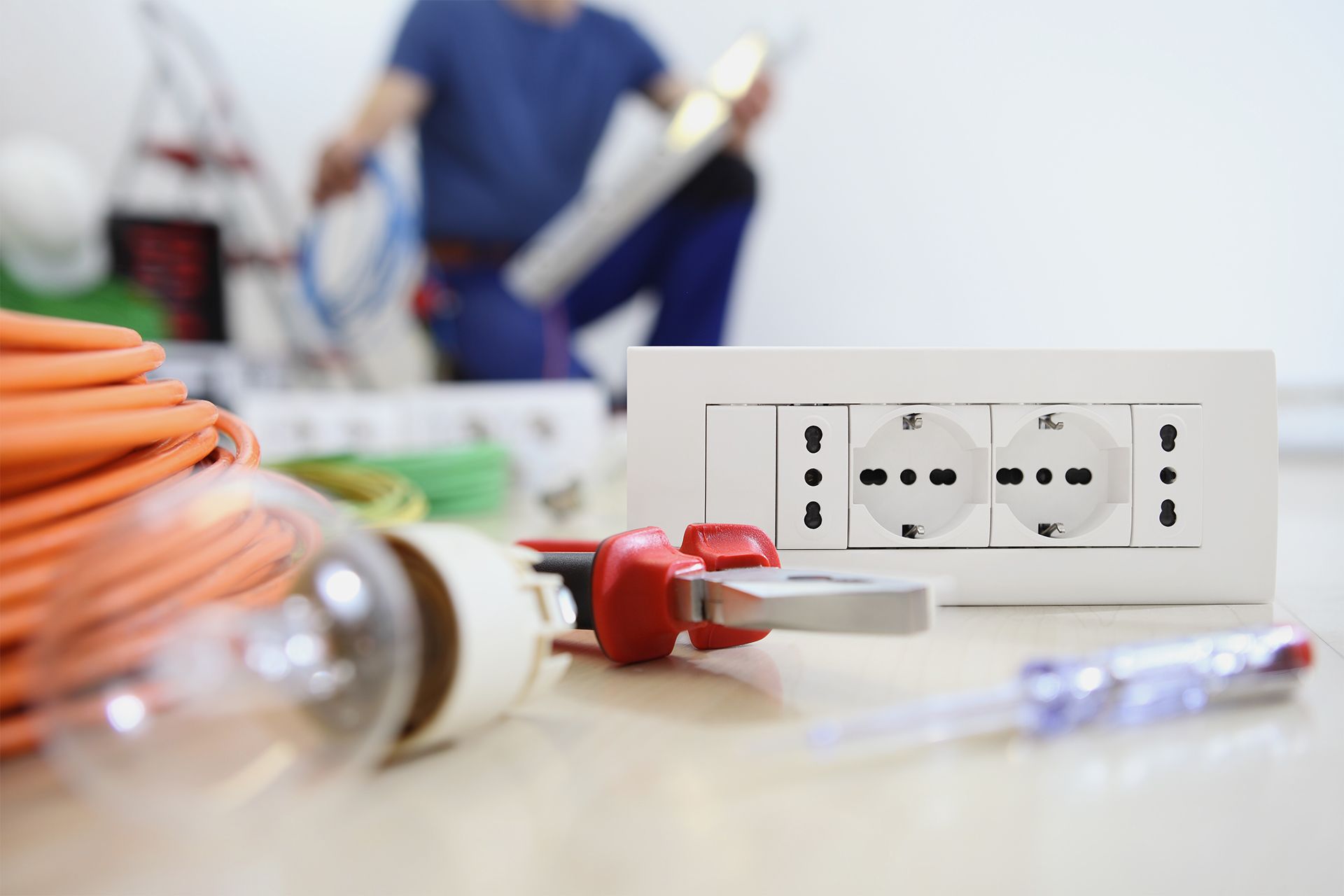 Electrician installing outlet; tools, wiring, and lightbulb in foreground.