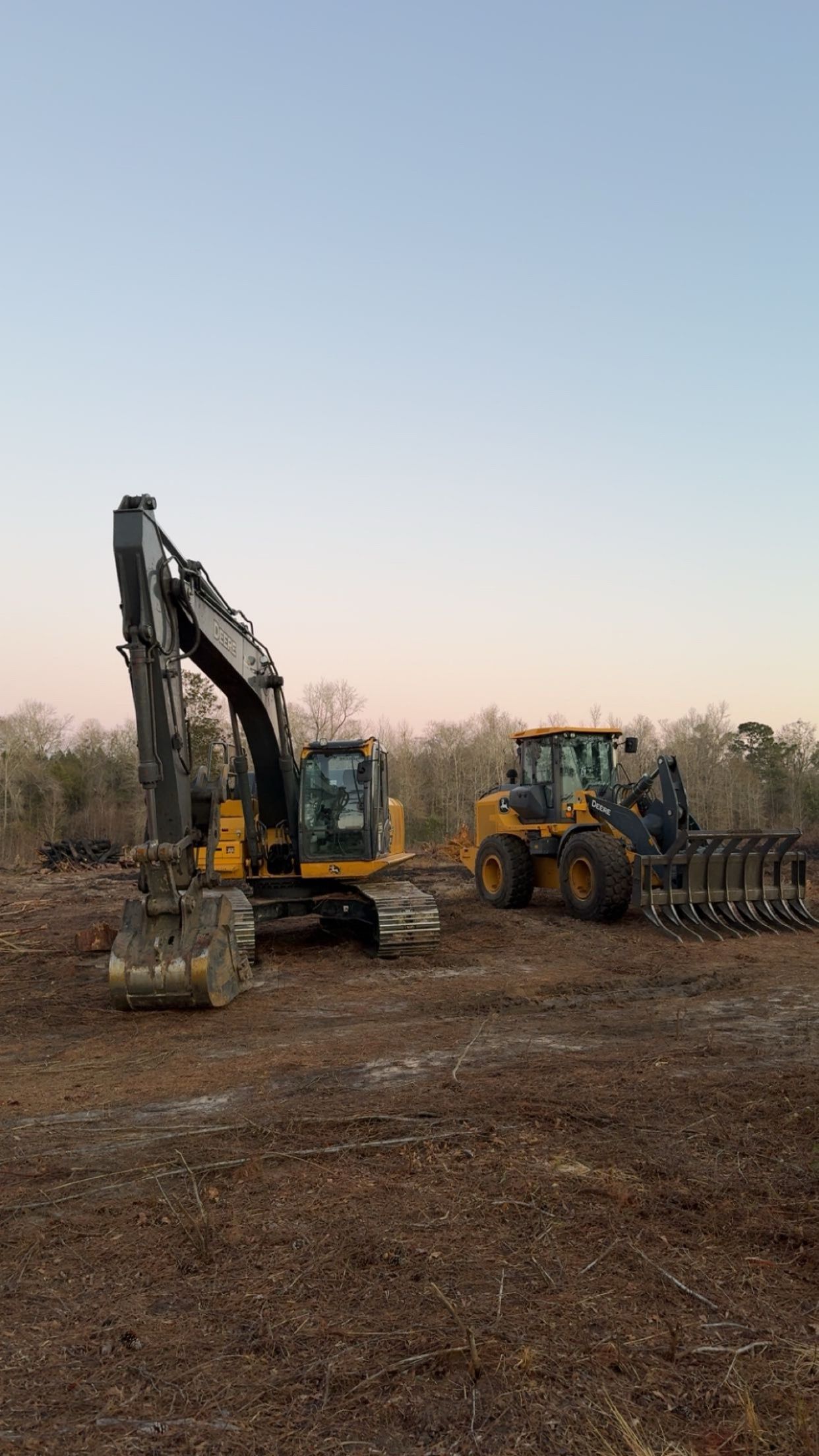 An excavator and a yellow tractor with a rake attachment parked on a cleared dirt construction site at dusk.