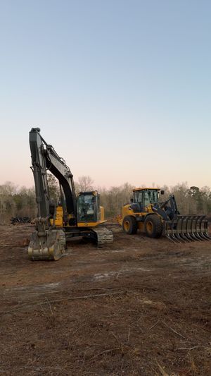 An excavator and a yellow tractor with a rake attachment parked on a cleared dirt construction site at dusk.