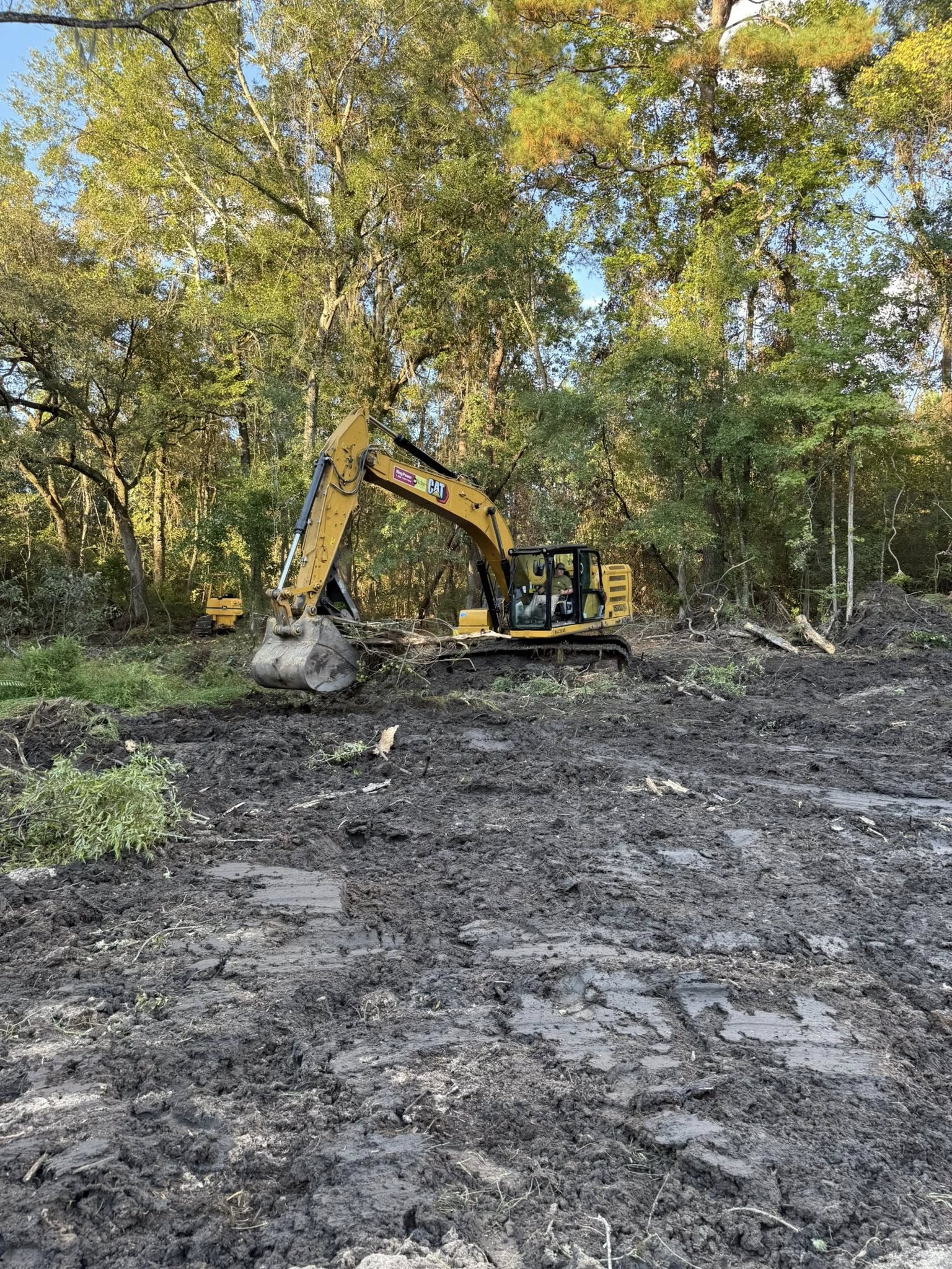 A yellow excavator sits in a muddy, cleared area surrounded by a lush, green forest.