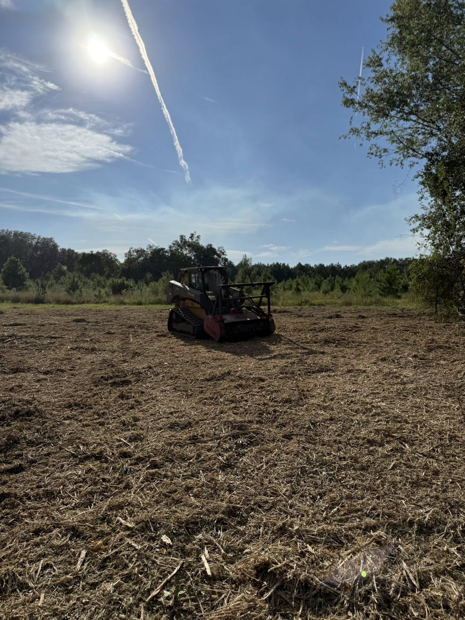 A construction skid steer clearing brush in a sunny, expansive field under a bright blue sky with a contrail.