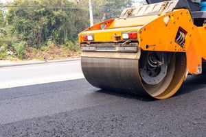 A yellow road roller compacts freshly laid dark asphalt on a road construction site.