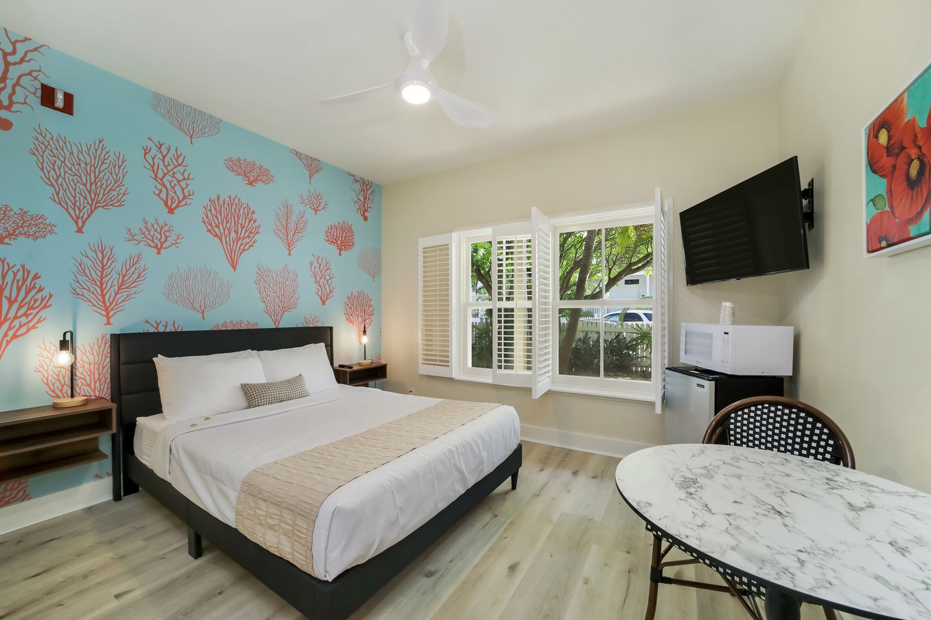 Hotel room with bed, table, TV, window, and coral-patterned wall.