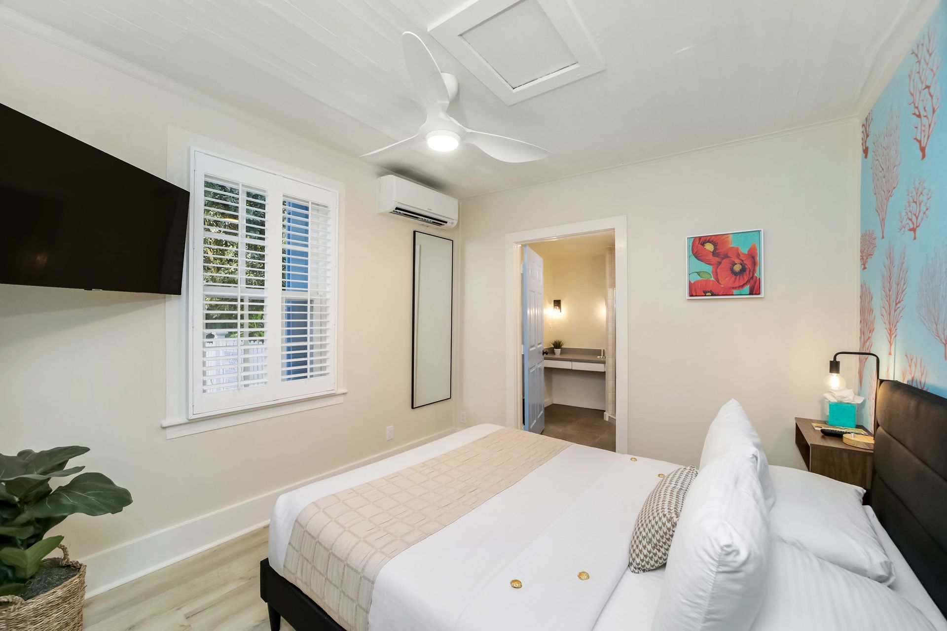 Bedroom with bed, TV, window, and small bathroom visible. Light walls and decor.