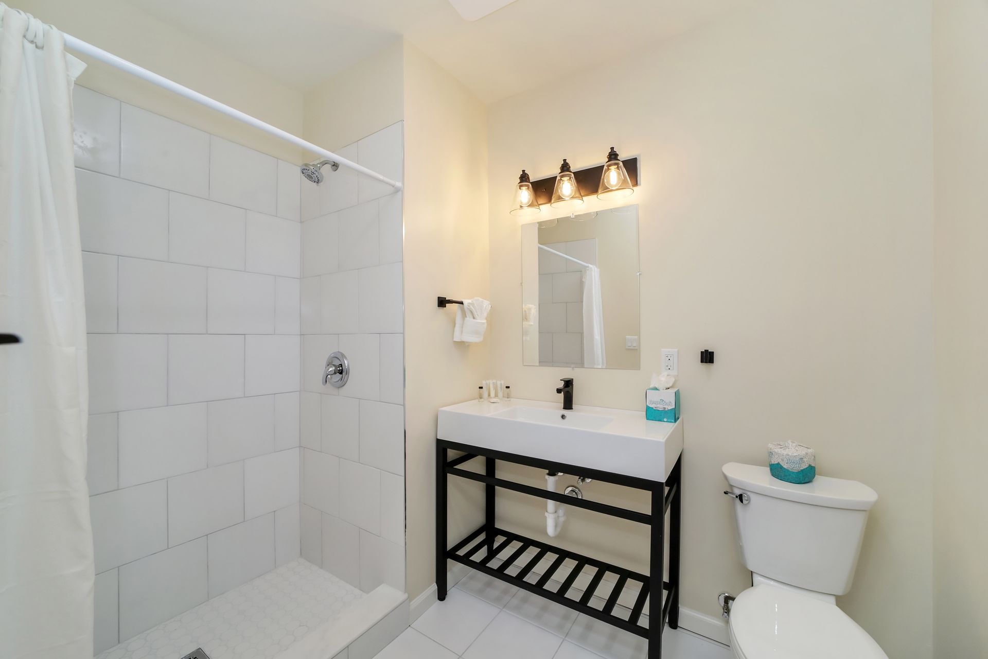 Bathroom with a white tiled shower, sink, toilet, and a black metal stand.