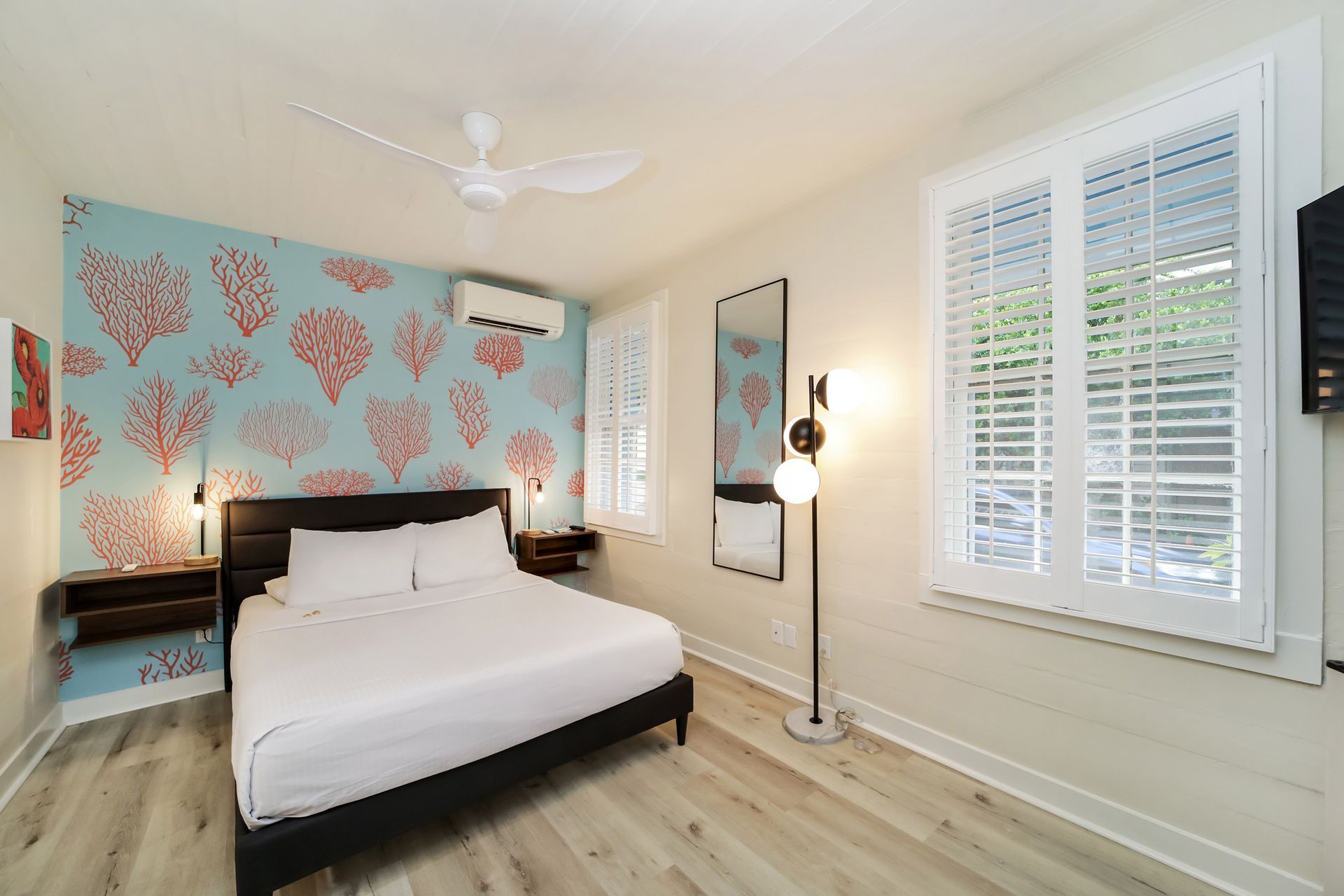 Bedroom with queen bed, coral patterned accent wall, white shutters, and a modern floor lamp.