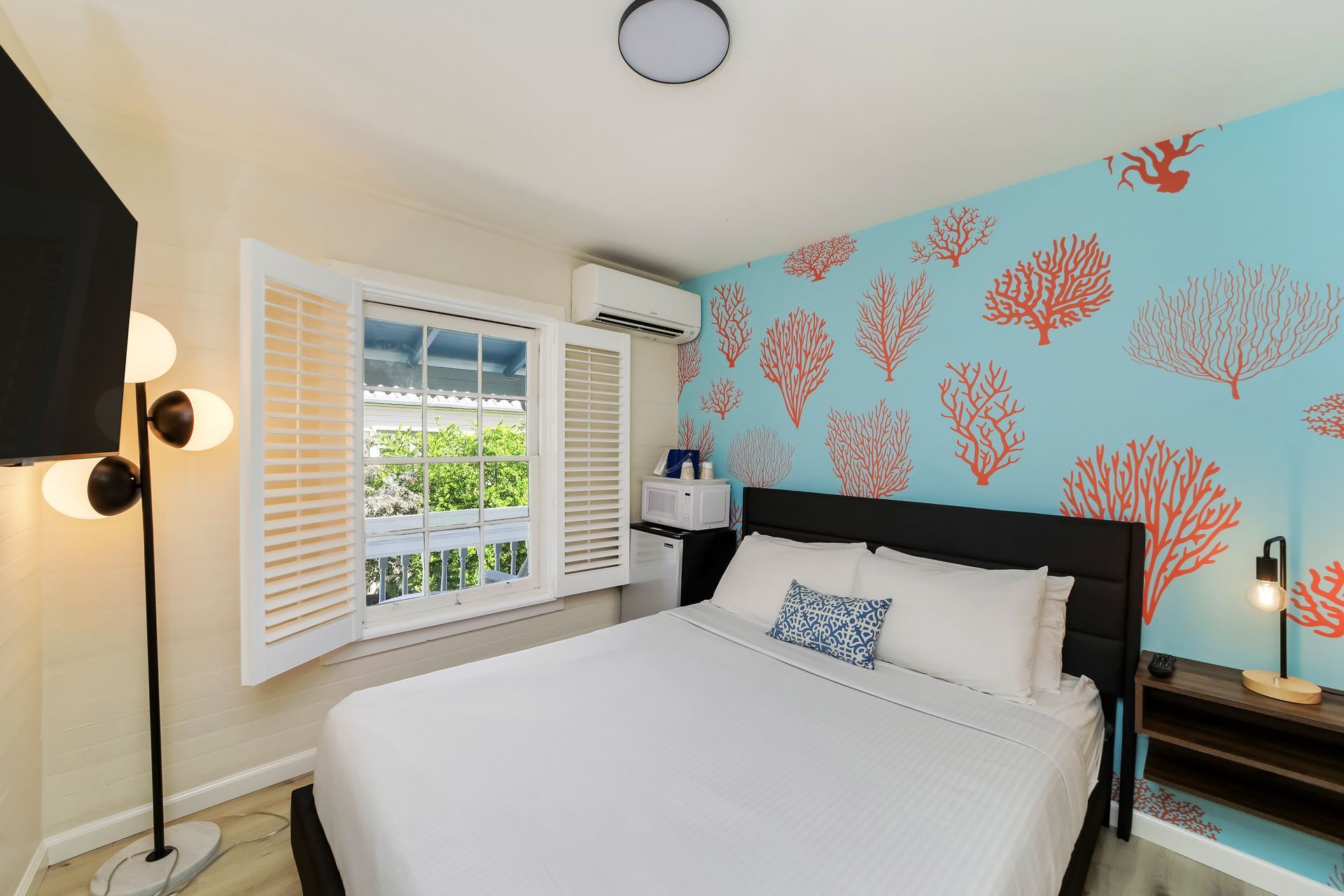 Bedroom with bed, coral wall, window with shutters, TV, and floor lamp.
