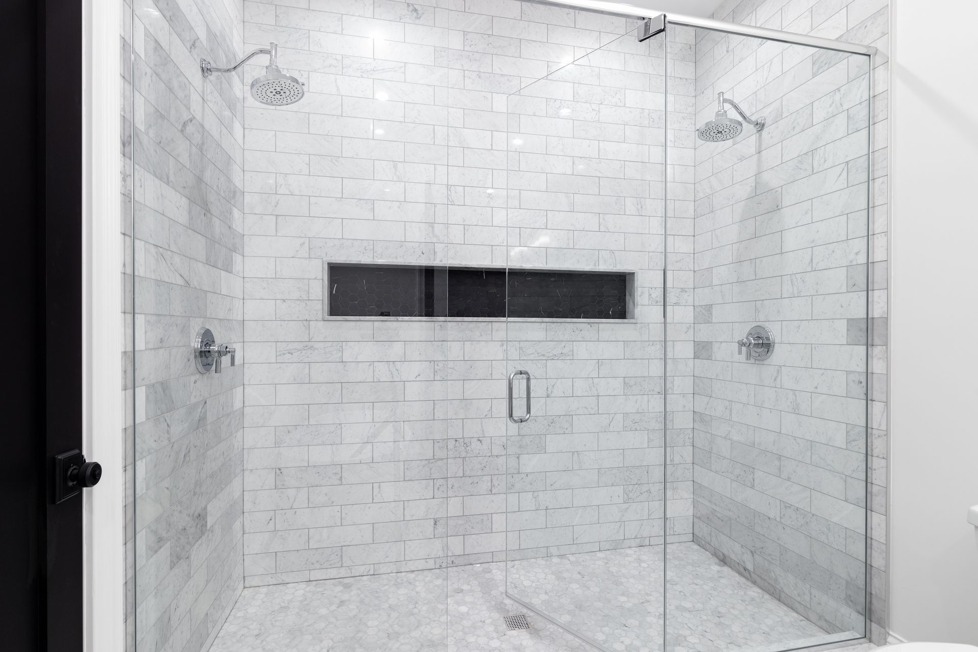 Glass-walled shower with white brick-like tiles, two showerheads, and a rectangular niche.