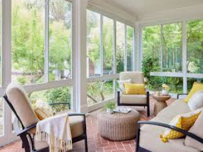 Sunroom with beige furniture, yellow pillows, and a brick floor, surrounded by windows with a green outdoor view.