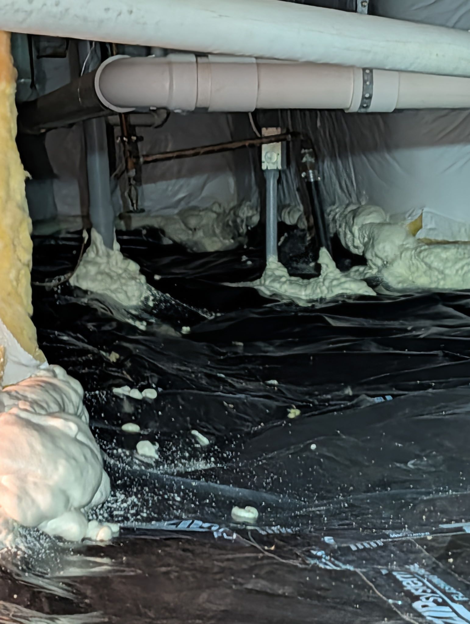 Insulated pipes and black plastic sheeting on a crawlspace floor with spray foam insulation around the edges