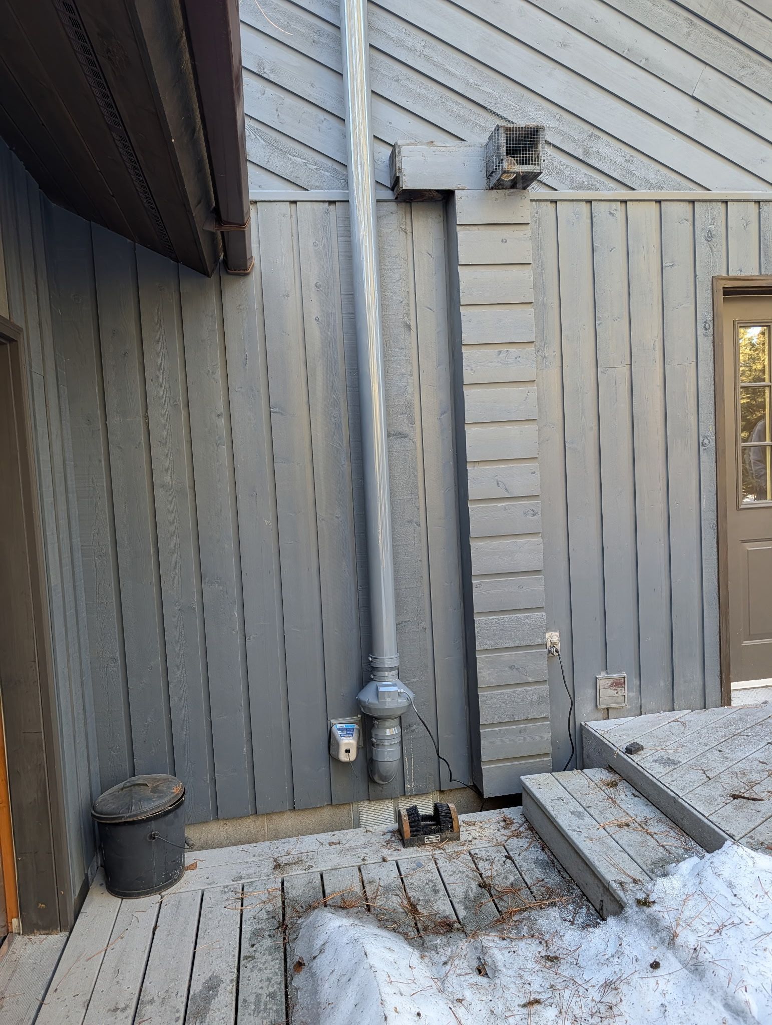 Gray outdoor wall with a vertical metal chimney pipe, vent box, and snow-covered ground beside a doorway