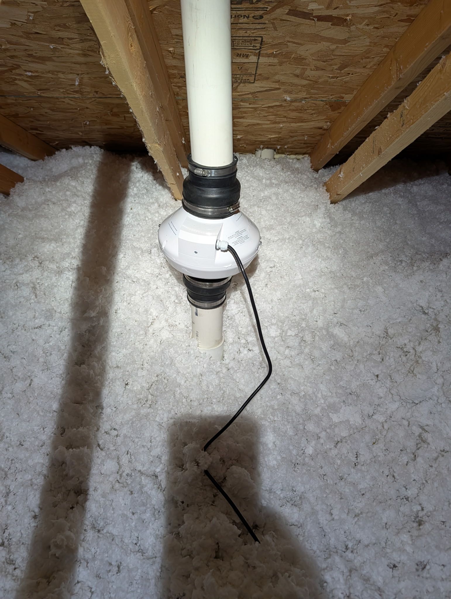White pipe and electrical device hanging in attic insulation beneath wooden rafters.