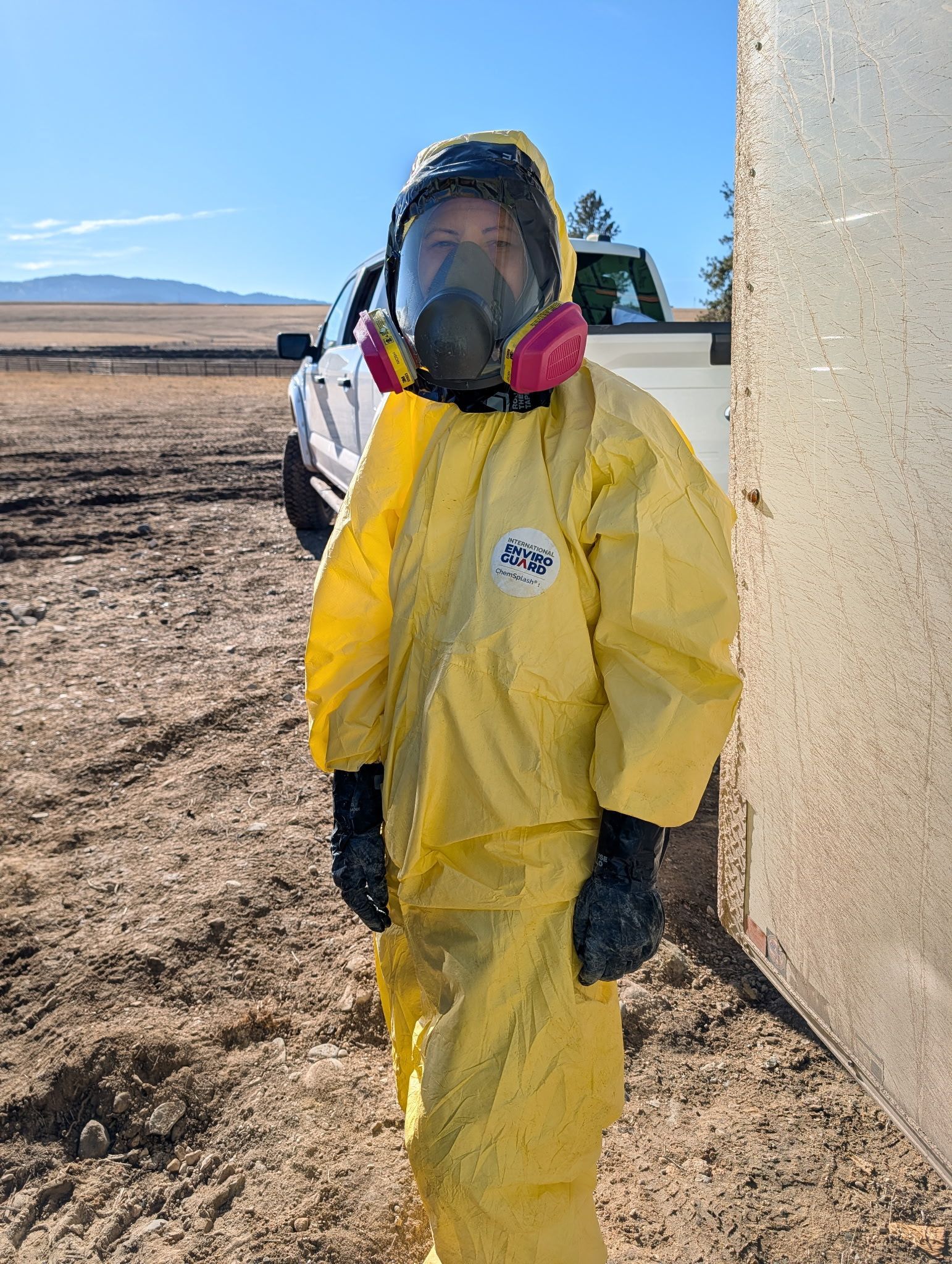 Person in yellow hazmat suit and respirator standing outdoors near a building in a snowy field.