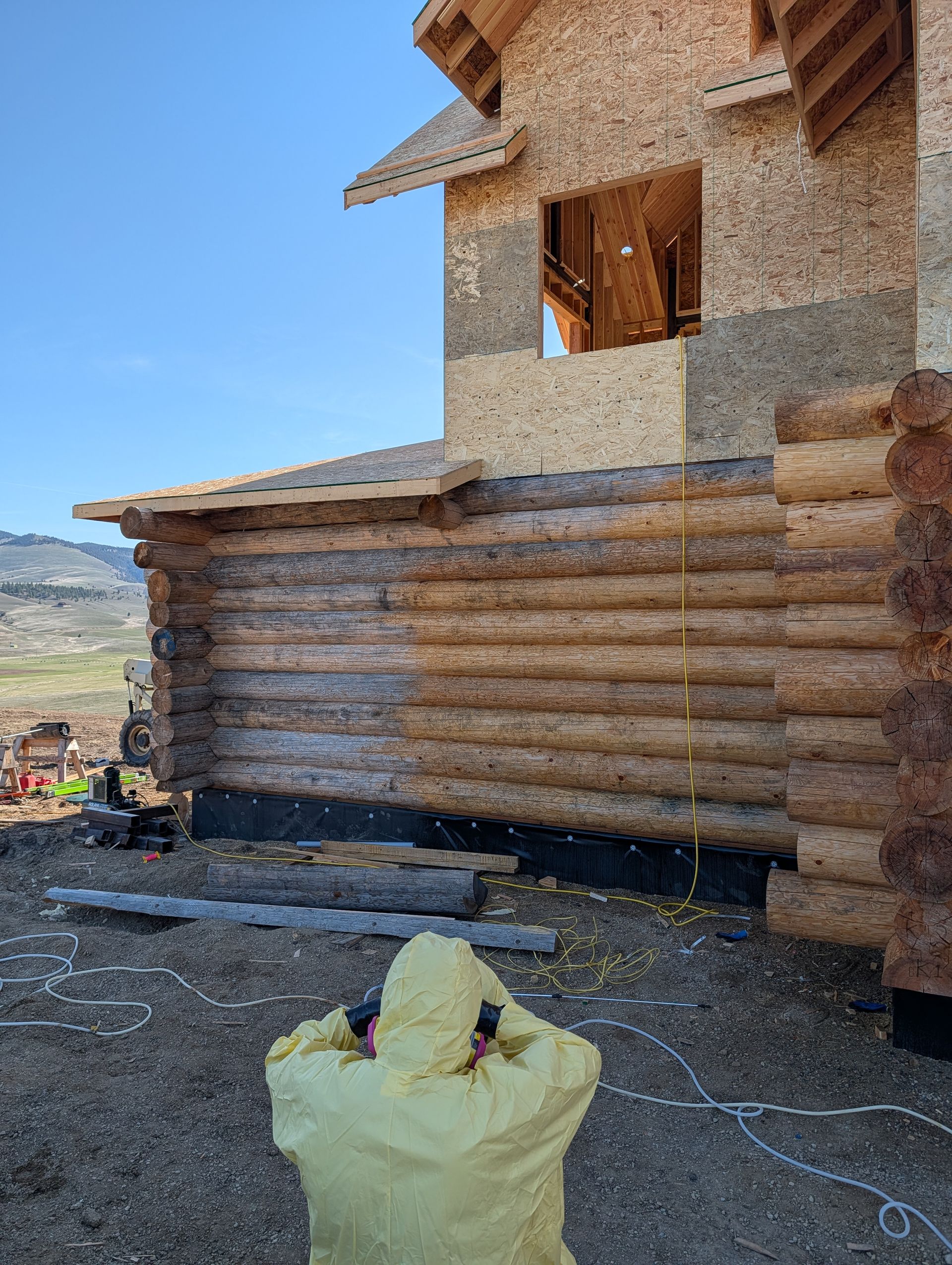 Wooden log cabin under construction in a dry landscape, with a worker in a yellow jacket in the foreground