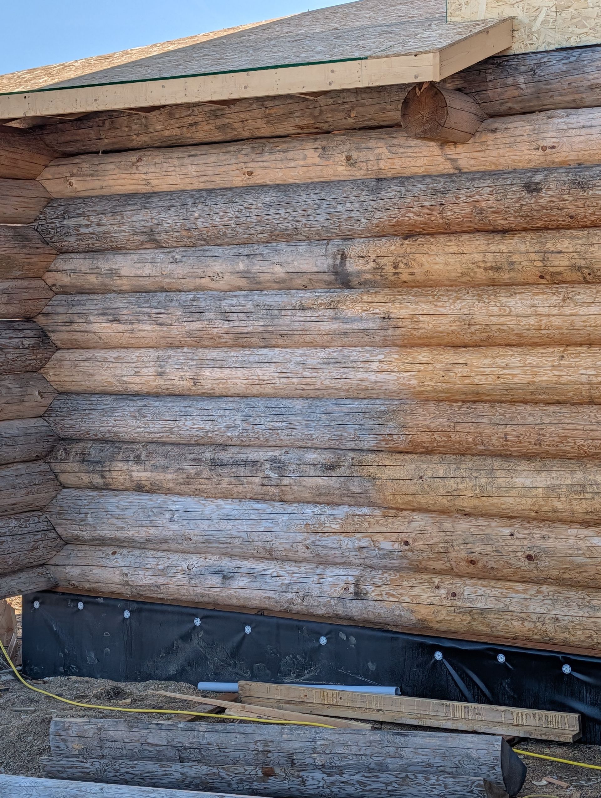 Weathered log wall under a roof eave, with black tarp at the base.