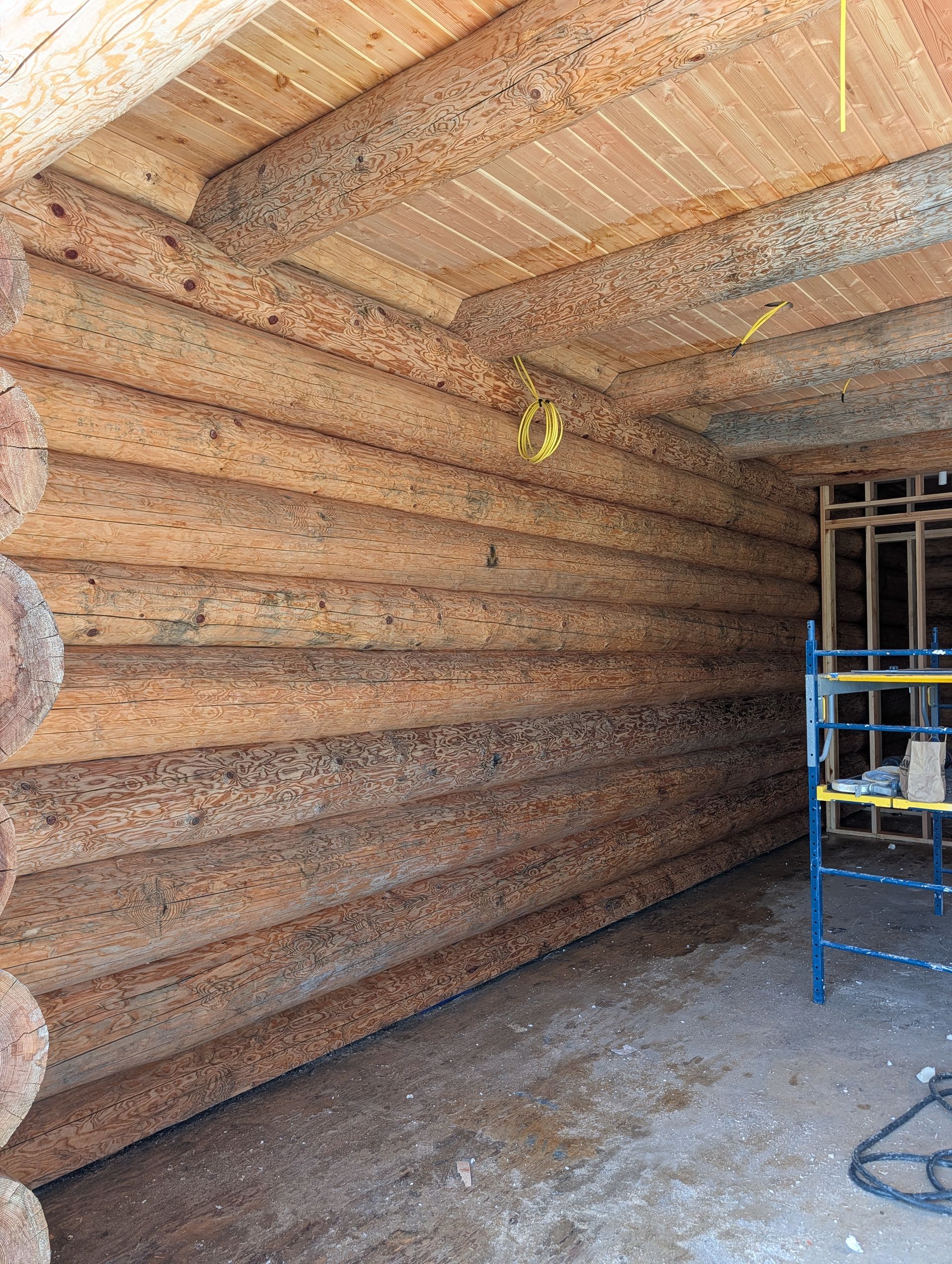 Rustic log interior with a wood floor, exposed ceiling, and blue scaffolding on the right.