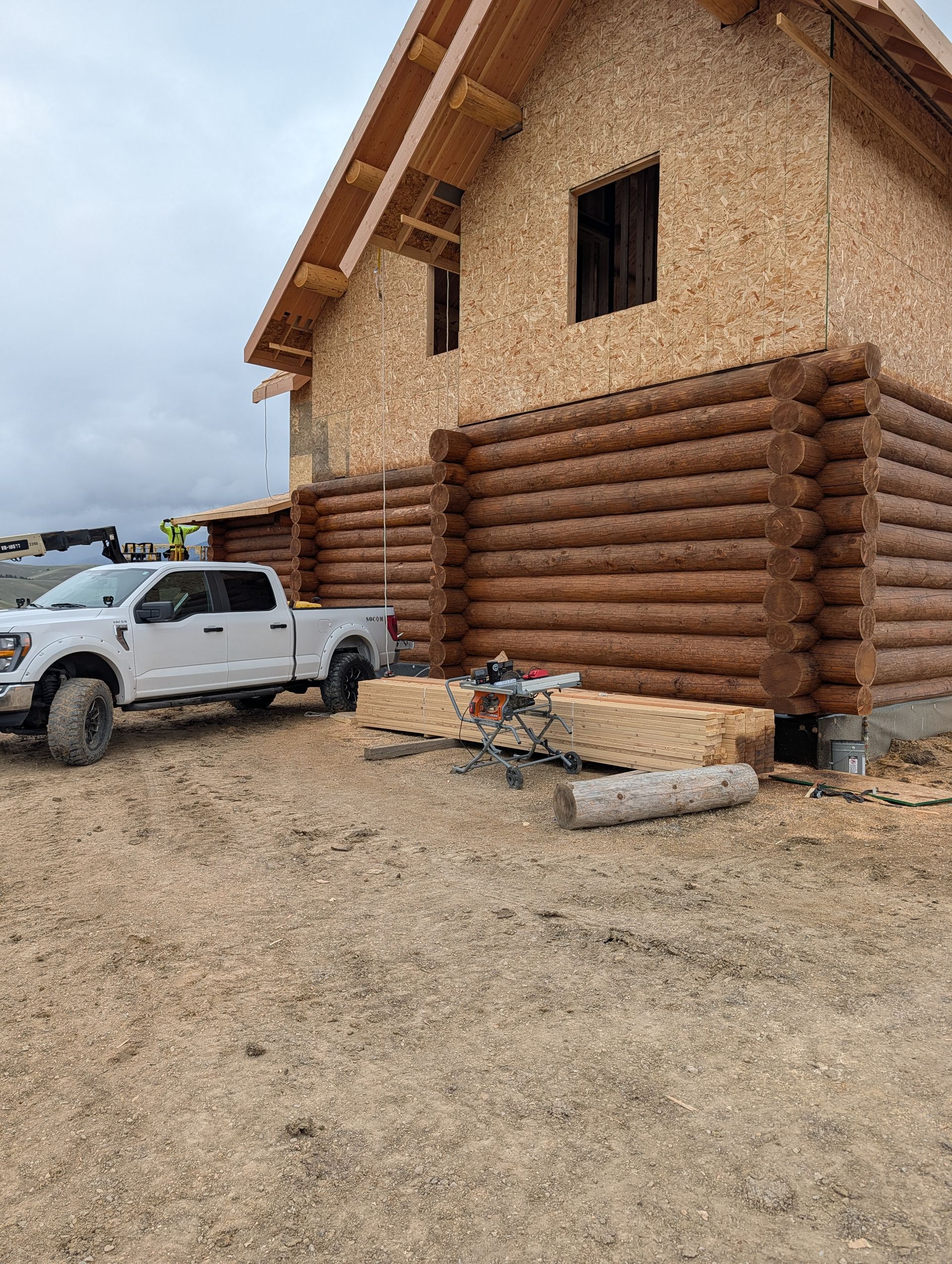 Stone-and-log house under construction beside a white pickup truck on a gravel lot.