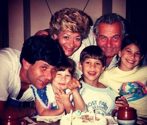 An old family photo featuring three generations gathered around a table. In front, Rebecca's dad leans forward. Next to him is Rebecca's younger sister (at age 3), brother (at age 7.5) and then herself (at age 9). Her grandparents embrace the children from behind. All are smiling and there is  a birthday cake in front of them. 