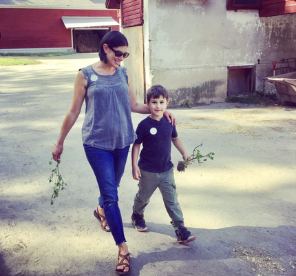 Rebecca walks with her young son on a dirt path. She wears sunglasses and smiles looking down with her arm on her son's shoulder. Her son smiles and looks directly at the camera. Both hold a small plant with visible roots.