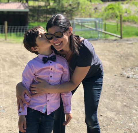 Rebecca embraces her young son outdoors on a sunny day. He is dressed in a pink button-up shirt with a navy bow tie, is leaning in to give her a kiss on the cheek. Rebecca is wearing sunglasses and smiles warmly while hugging him close. The background features a dirt path and green trees.