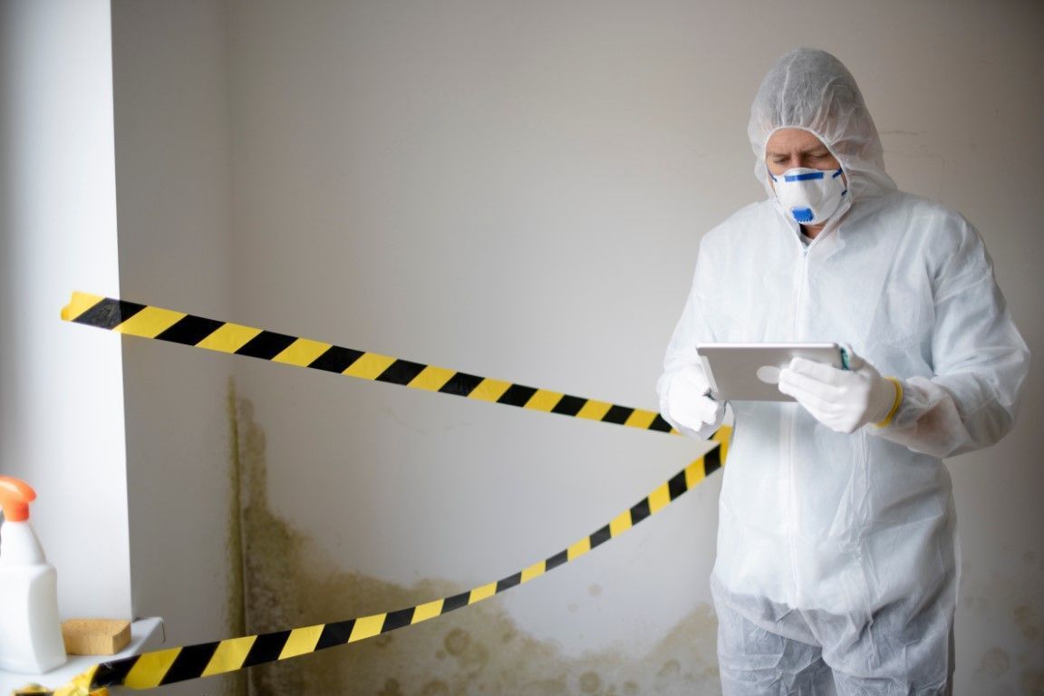 Worker in white protective suit inspecting mold-damaged wall behind caution tape