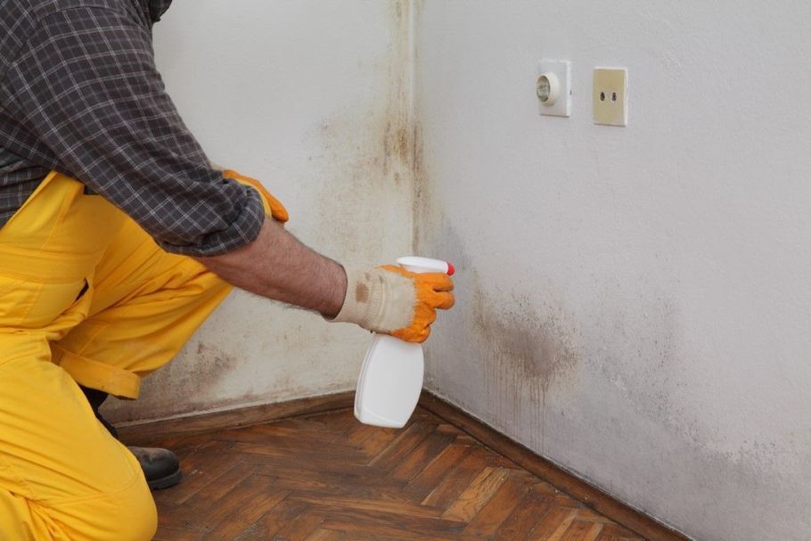 Worker spraying cleaner along a dirty wall corner above a wood floor
