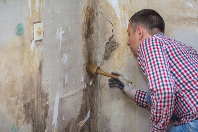 Man in a red plaid shirt scraping peeling paint from a wall with a scraper
